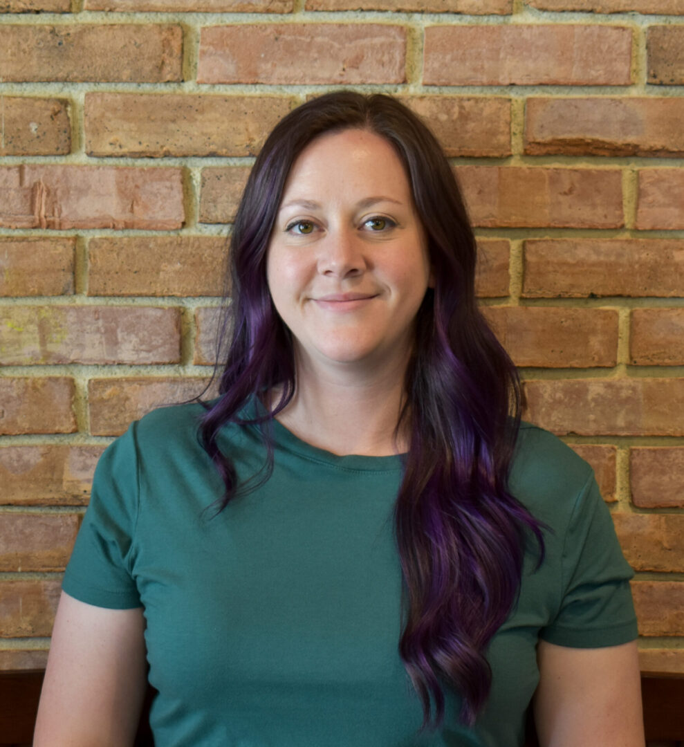 A woman with purple hair standing in front of a brick wall.